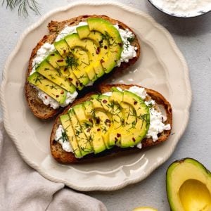 Plate with two pieces of avocado cottage cheese toast on a light grey backdrop surrounded by ingredients