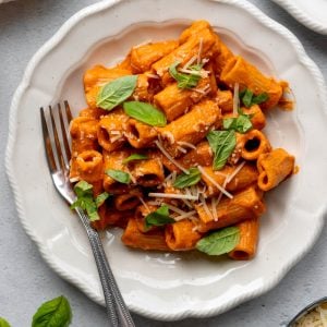 white plate with creamy tomato sauce rigatoni topped with basil leaves and parmesan cheese, with a fork
