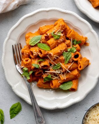 white plate with creamy tomato sauce rigatoni topped with basil leaves and parmesan cheese, with a fork