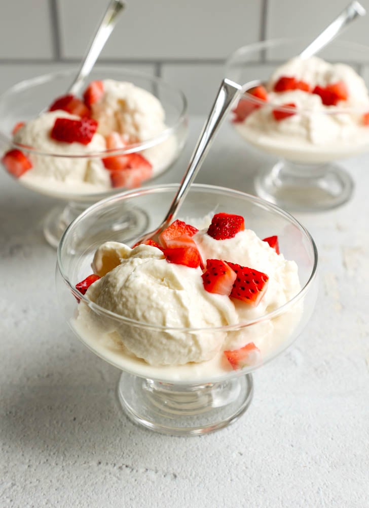 three ice cream bowls with scoops of vanilla greek yogurt topped with chopped strawberries, with small spoons