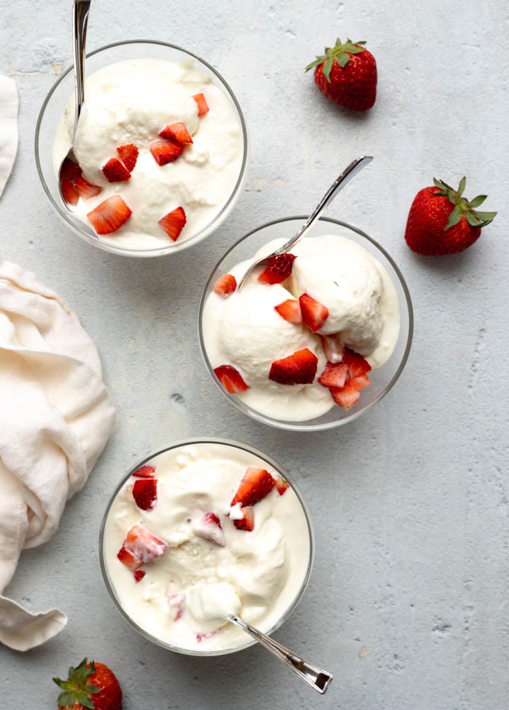 three ice cream bowls filled with vanilla greek yogurt and chopped strawberries with silver spoons, on a light grey backdrop