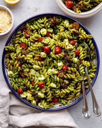 serving bowl of tri color pasta salad surrounded by ingredients with two serving spoons in the bowl on a light grey backdrop