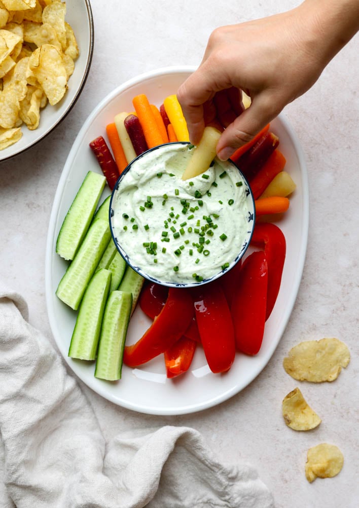 hand dipping into a small bowl of veggie dip surrounded by cucumbers, bell pepper, and carrots