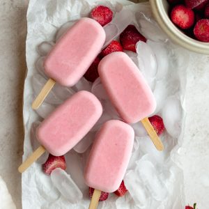 4 strawberry popsicles on a plate with ice, surrounded by a bowl of frozen strawberries and a few fresh strawberries