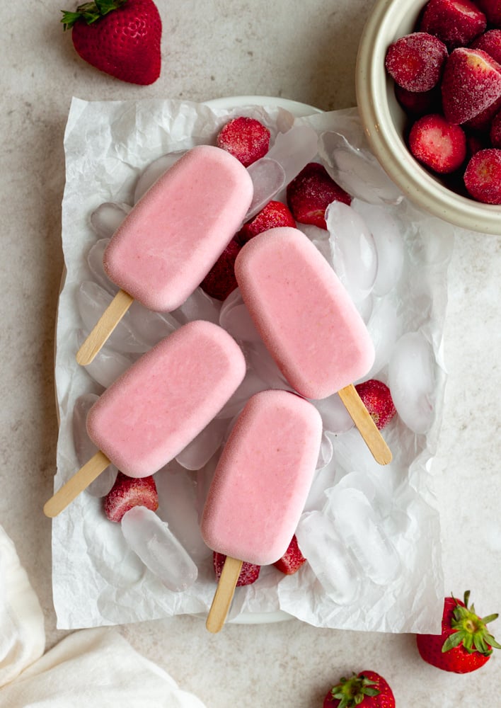 4 strawberry popsicles on a plate with ice, surrounded by a bowl of frozen strawberries and a few fresh strawberries
