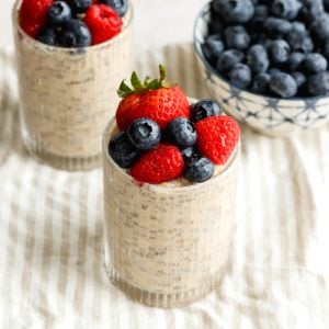 Two cups of overnight oats topped with berries, and a bowl of blueberries on a light striped dish towel