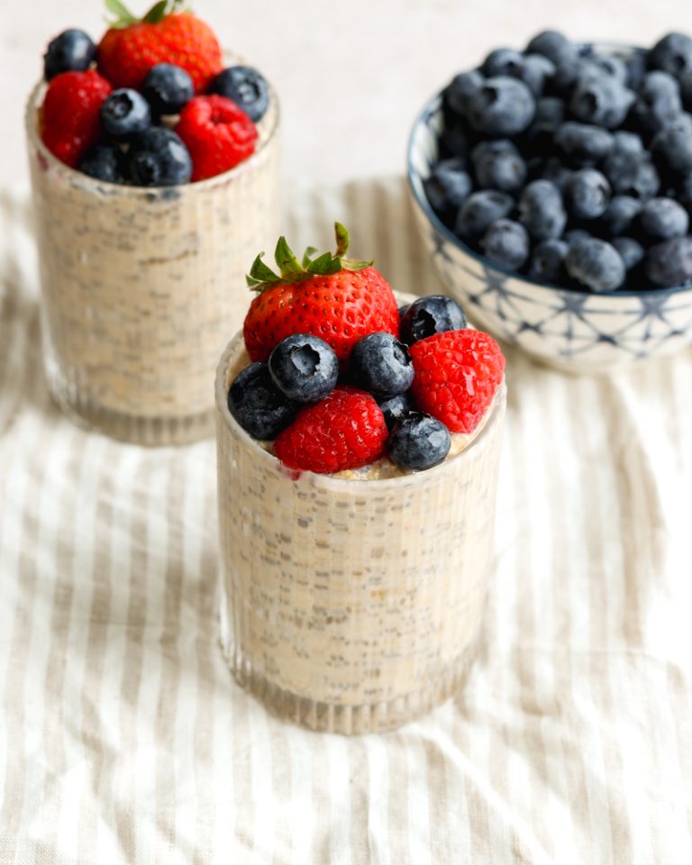 Two cups of overnight oats topped with berries, and a bowl of blueberries on a light striped dish towel