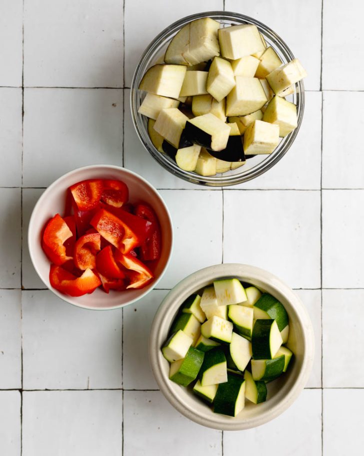 cubed eggplant, zucchini, and bell pepper in bowls on a white tile backdrop