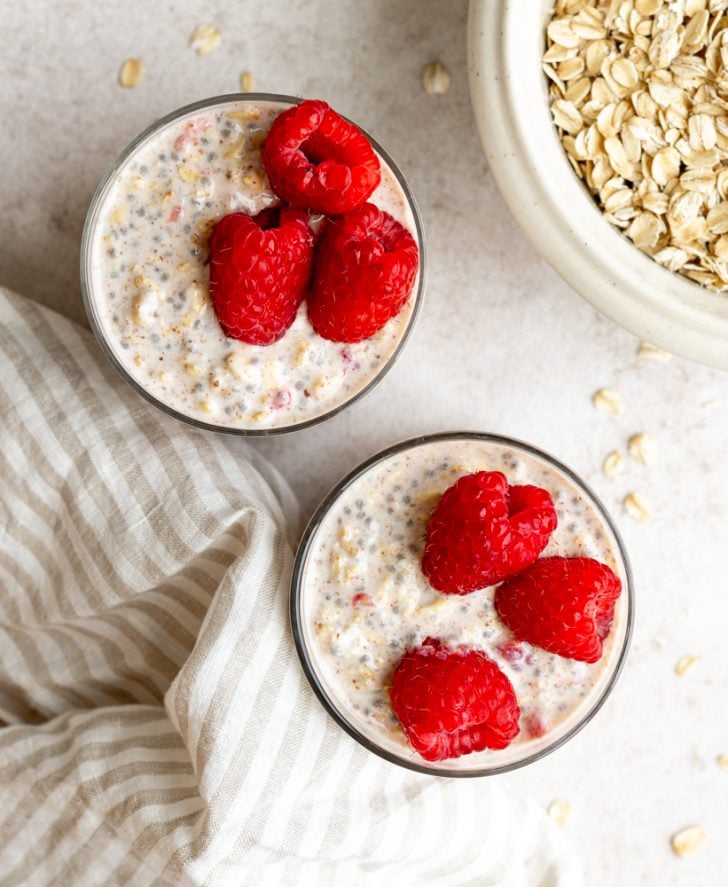 two glasses of overnight oats with raspberries from above next to a bowl of raw oats