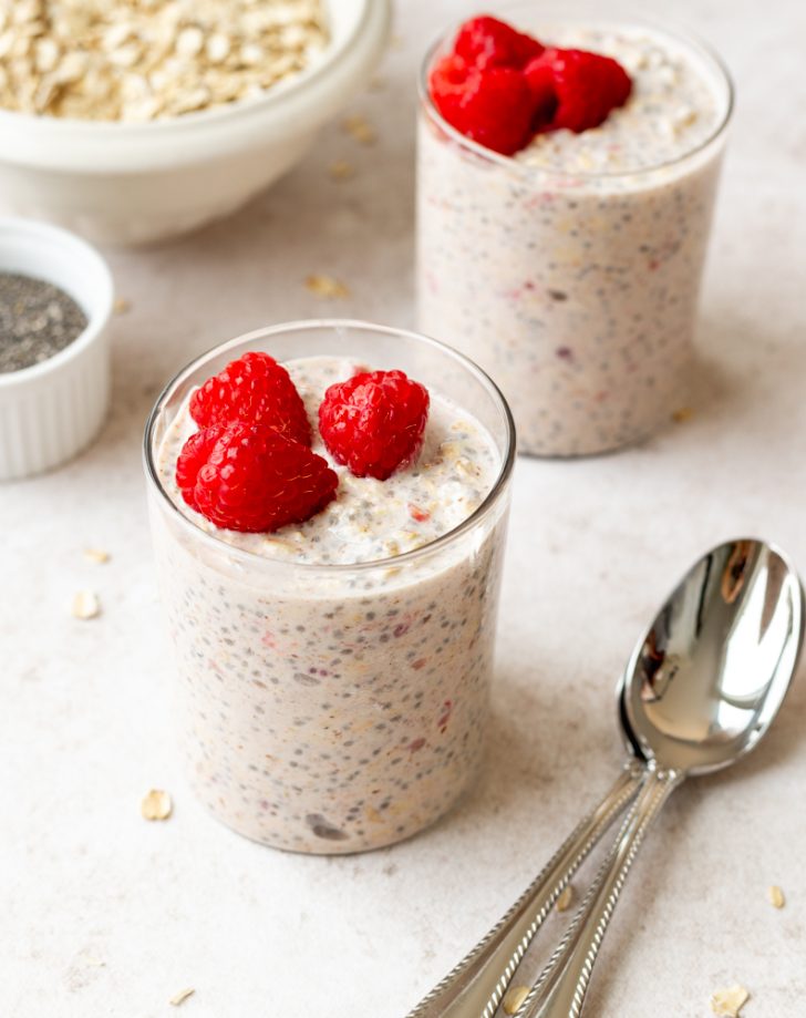 two glass cups of overnight oats with raspberries and silver spoons in the foreground, bowls of oats and chia seeds in the background