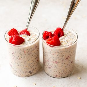 two glass cups of overnight oats with raspberries and silver spoons