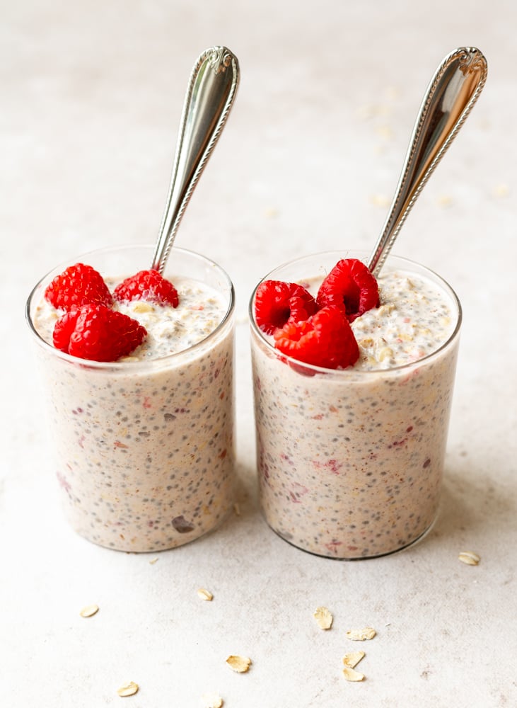 two glass cups of overnight oats with raspberries and silver spoons