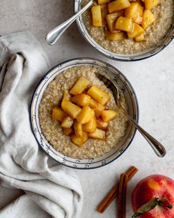 two bowls of apple cinnamon steel cut oatmeal with silver spoons, an apple and cinnamon sticks next to the bowls