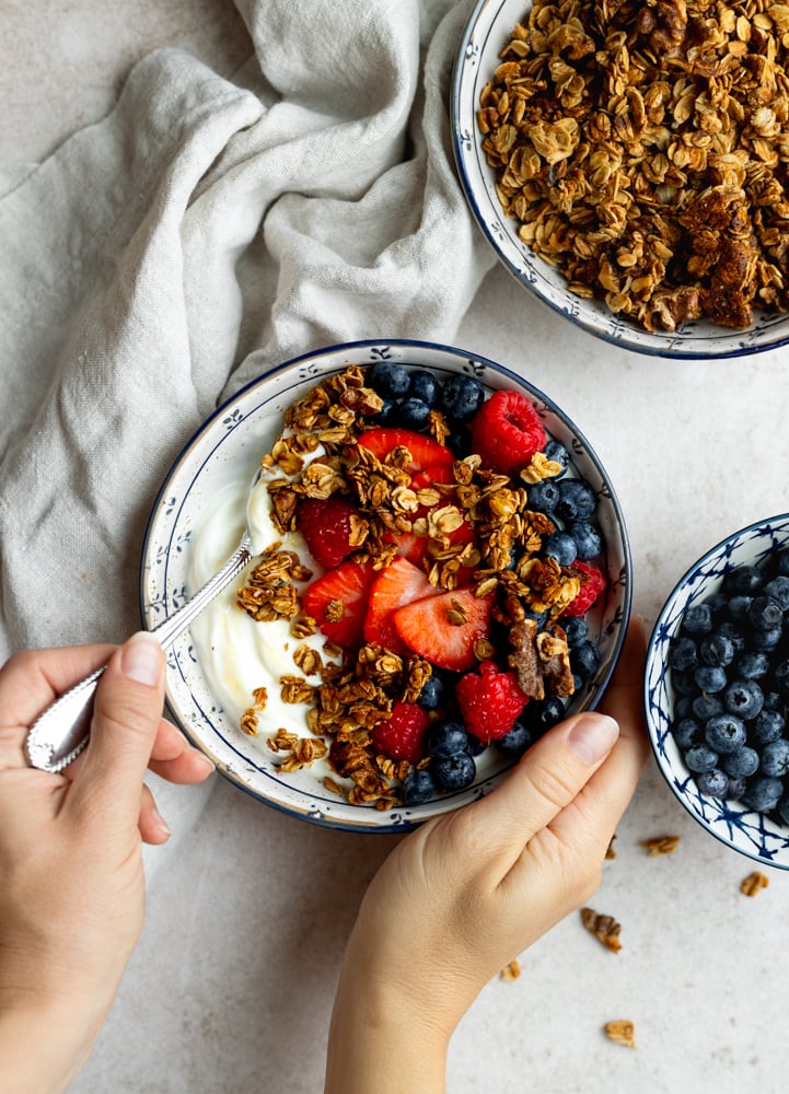 hand scooping into a yogurt granola fruit bowl surrounded by a bowl of granola and blueberries