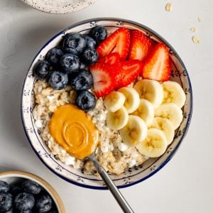 bowl of cottage cheese oatmeal with fresh fruit and a spoonful of peanut butter surrounded by small bowls of different ingredients