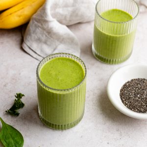 two small glasses with spinach kale smoothie next to a small dish of chia seeds, spinach leaves, and bananas
