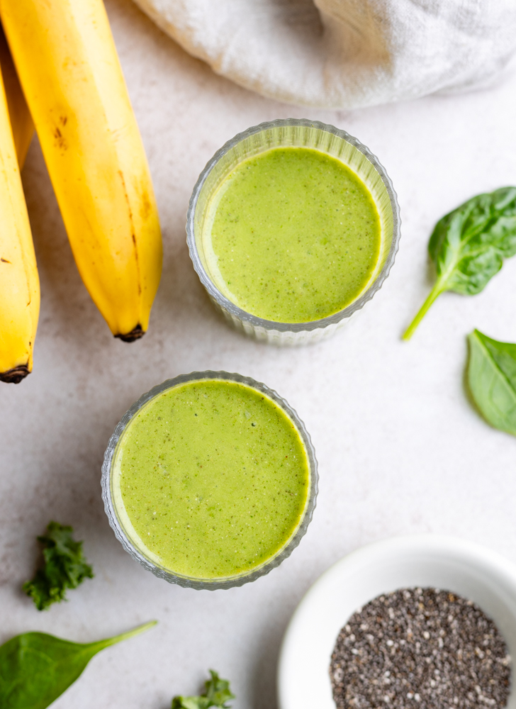 two small glasses with spinach kale smoothie next to a small dish of chia seeds, spinach leaves, and bananas