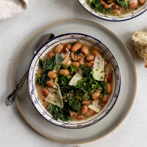 ceramic bowl with brothy beans and greens, topped with parmesan cheese on a plate with a spoon