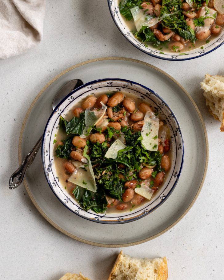 ceramic bowl with brothy beans and greens, topped with parmesan cheese on a plate with a spoon