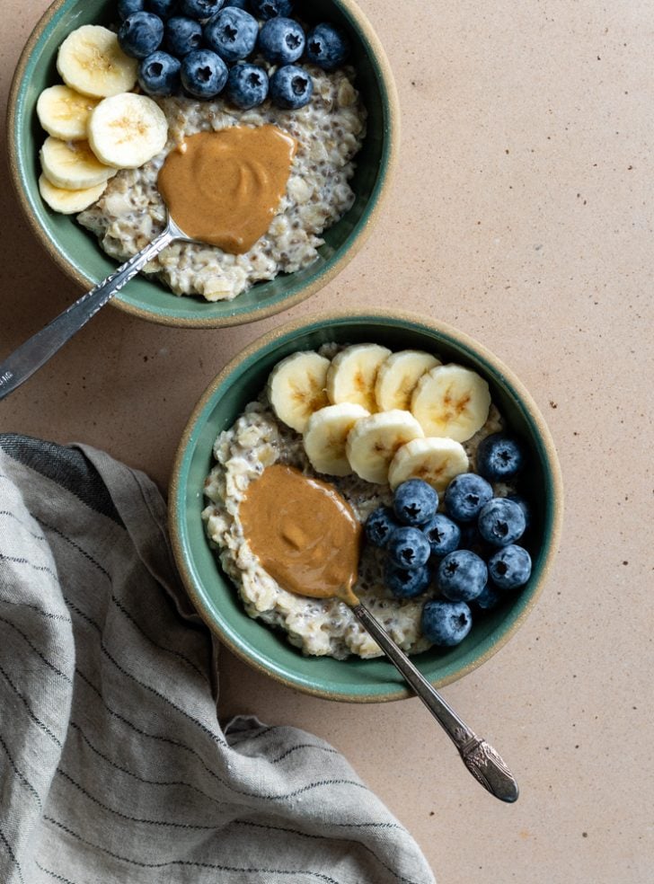 two bowls of greek yogurt oatmeal with banana slices, blueberries, and a spoon of peanut butter