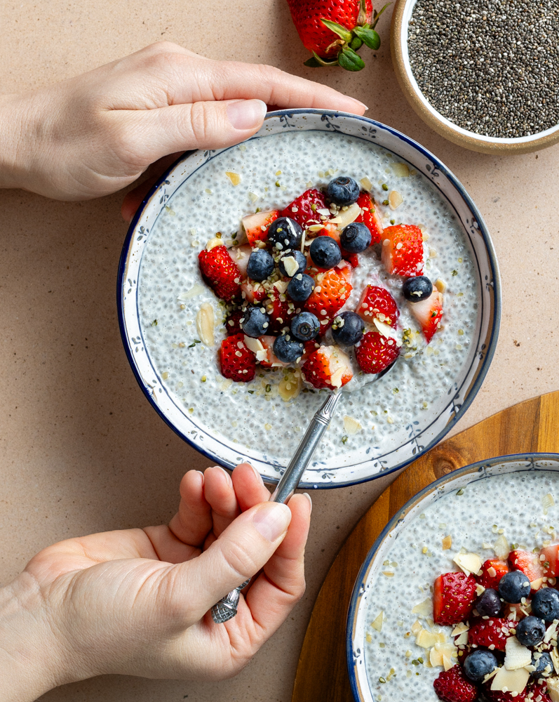 hands taking a spoonful of chia pudding from a blue and white bowl with chia pudding topped with fresh berries and sliced almonds