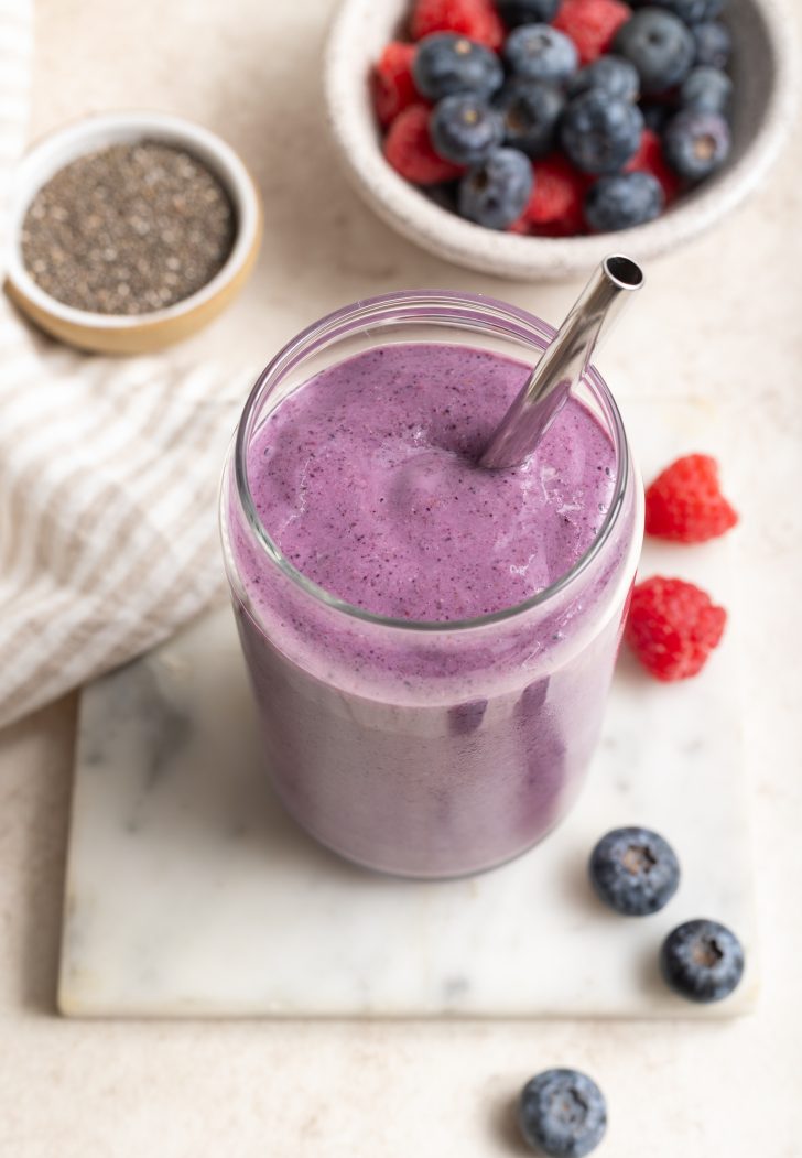 high fiber smoothie in a glass jar with a silver metal spoon on a white marble slab, surrounded by little bowl of chia seeds and some berries