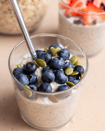 glass cup with overnight oats topped with blueberries and pepitas with a stainless steel spoon